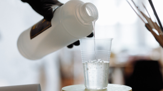 a hand wearing black glove pouring clear resin from a bottle into a small clear cup